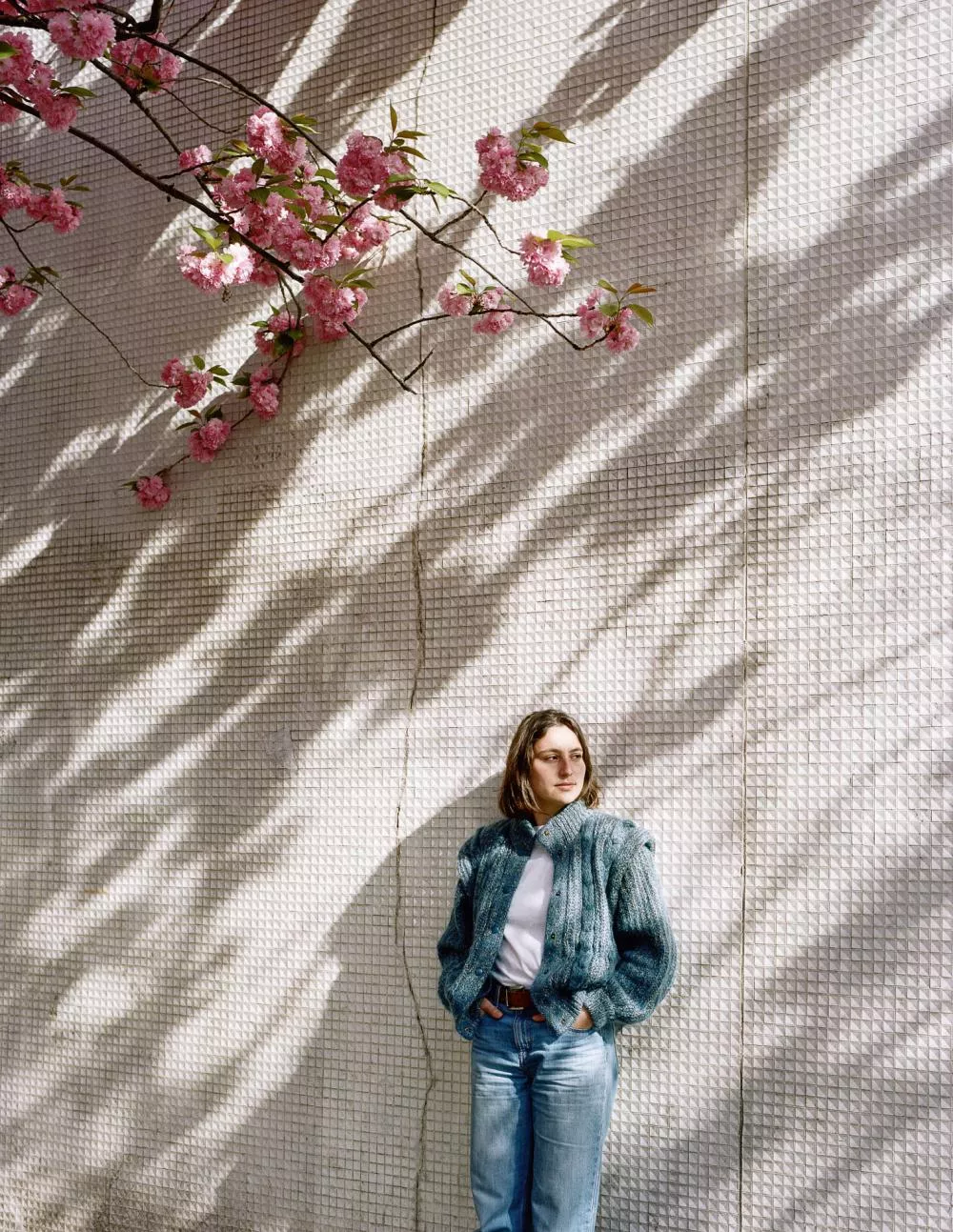 Une personne se tient contre un mur texturé, sous des branches de fleurs roses. Elle porte un pull bleu et un jean, avec les mains dans les poches, et est éclairée par la lumière du soleil, créant des ombres sur le mur.