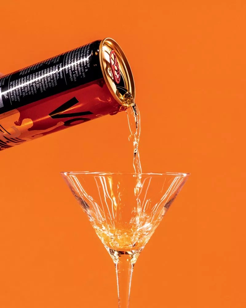 Une canette est en train de verser une boisson dans un verre à cocktail sur un fond orange.