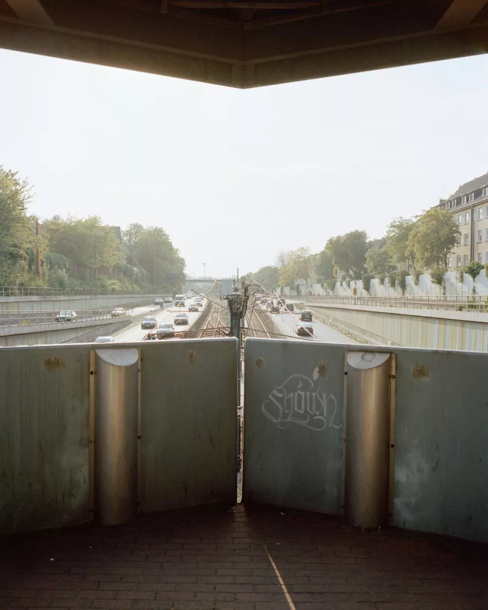 Une vue depuis un pont surplombant une autoroute avec plusieurs voitures en circulation. Des voies ferrées se trouvent au centre, entourées de barrières métalliques. Des arbres et des bâtiments résidentiels bordent la route.