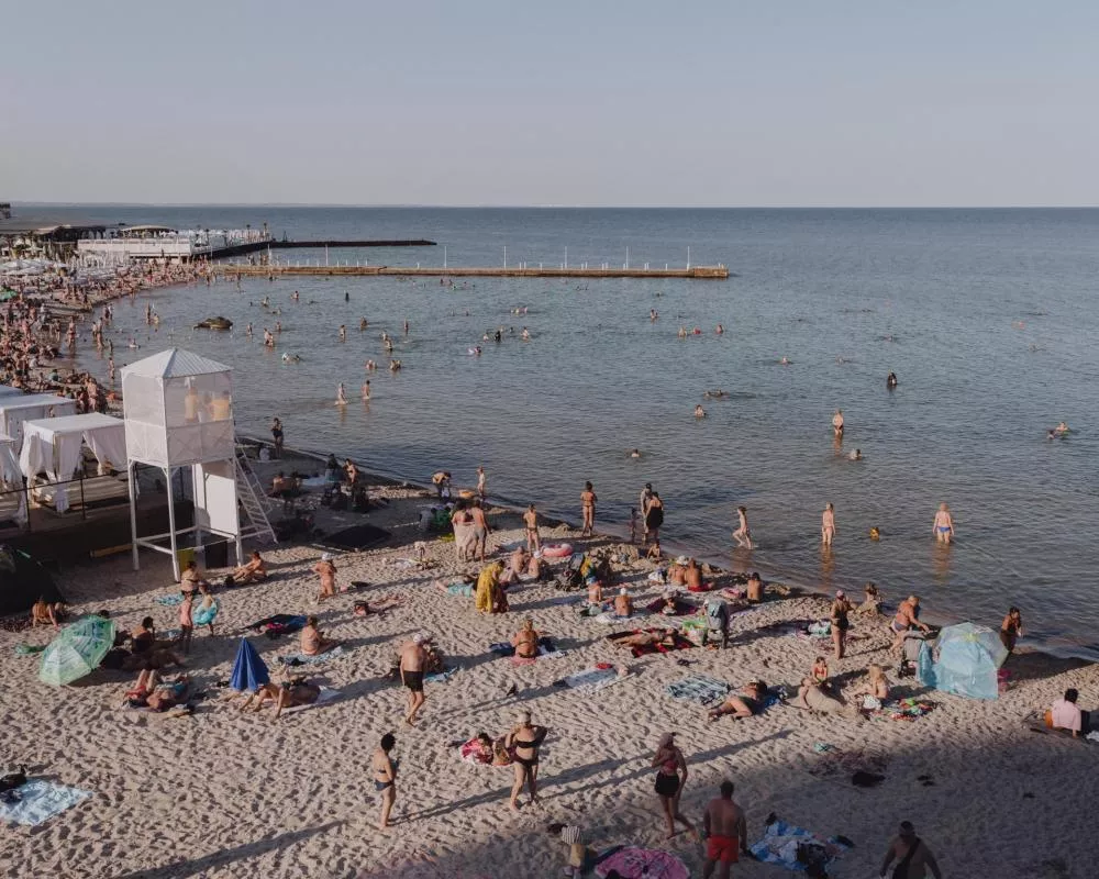 Une plage animée avec de nombreuses personnes se relaxant sur le sable et se baignant dans la mer. On voit des parasols, des serviettes et une tour de surveillance blanche sur la plage.