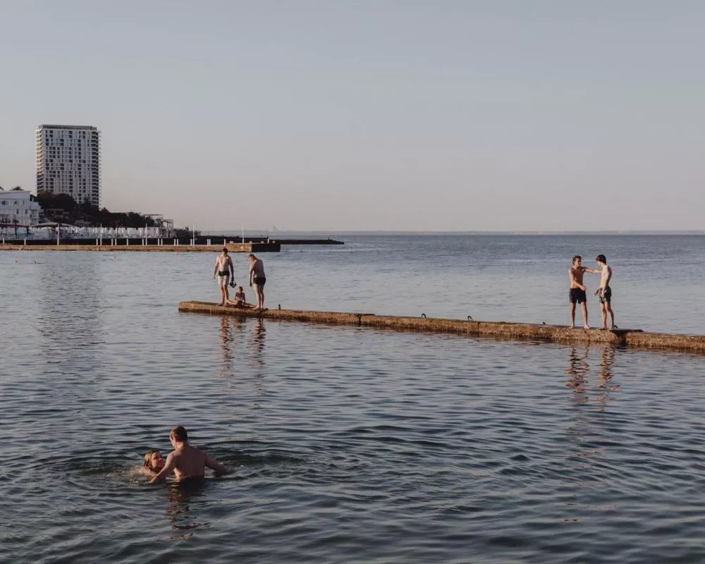 Des personnes profitent d'une journée ensoleillée au bord de la mer. Deux personnes sont dans l'eau et trois autres se trouvent sur un quai, discutant ou se préparant à entrer dans l'eau. En arrière-plan, on aperçoit des bâtiments et un grand immeuble.