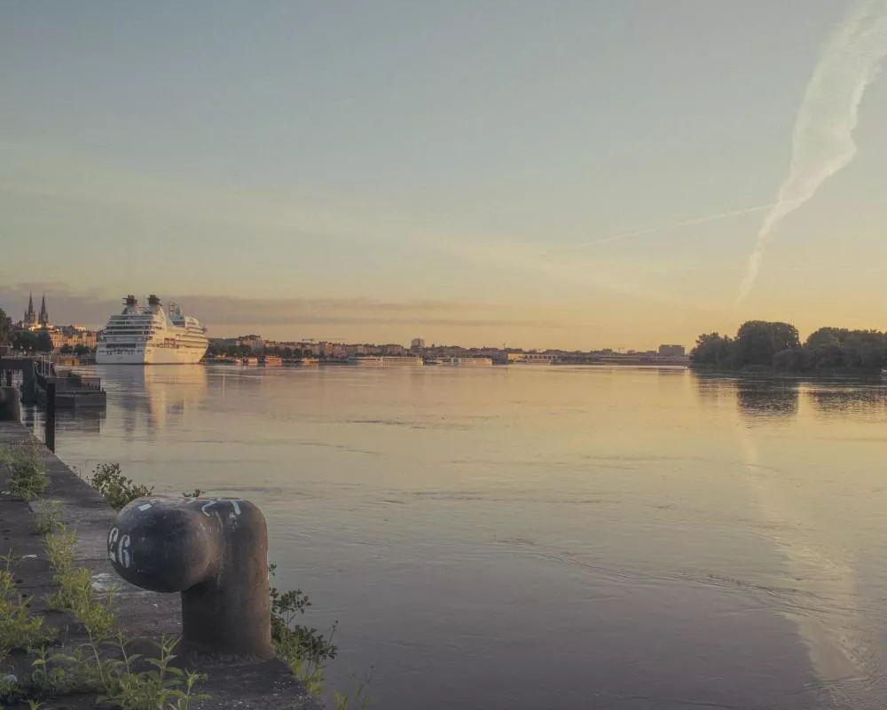 Un grand navire de croisière est amarré le long d'une rive, avec un ciel dégagé et des reflets dans l'eau. Des bâtiments et des arbres sont visibles à l'arrière-plan, sous une lumière douce de fin de journée.