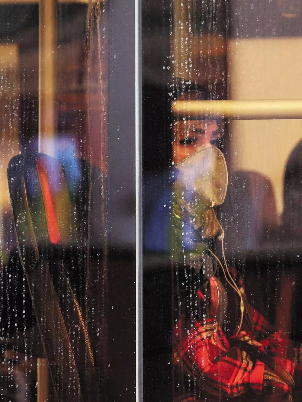 Une personne portant un masque est assise à l'intérieur d'un bus, regardant à travers une fenêtre couverte de gouttes de pluie.