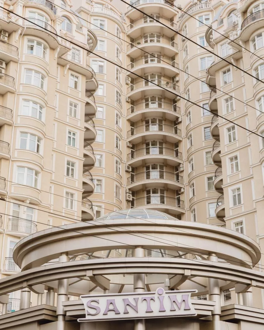 Un grand immeuble résidentiel beige avec des balcons arrondis, devant lequel se trouve une structure moderne avec le mot "SANTIM" affiché.
