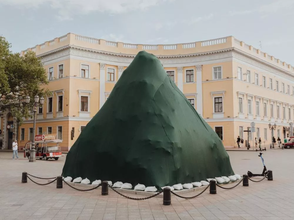 Un grand objet en forme de cône est recouvert d'une bâche verte, entouré de sacs de sable, situé sur une place pavée devant un bâtiment jaune avec des fenêtres et des moulures blanches.