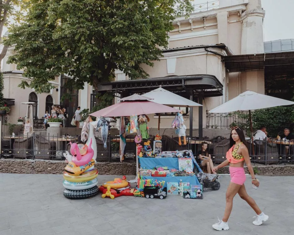 Une femme marche devant un stand de rue coloré sous des parasols, proposant des jouets gonflables et divers articles. Des personnes sont assises en arrière-plan à une terrasse, près d'un bâtiment ancien et d'un arbre.