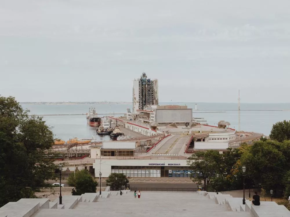 Une vue depuis le haut d'un escalier surplombant un port avec des bateaux amarrés, des bâtiments modernes et la mer à l'horizon. Des arbres encadrent la scène et quelques personnes descendent les marches.