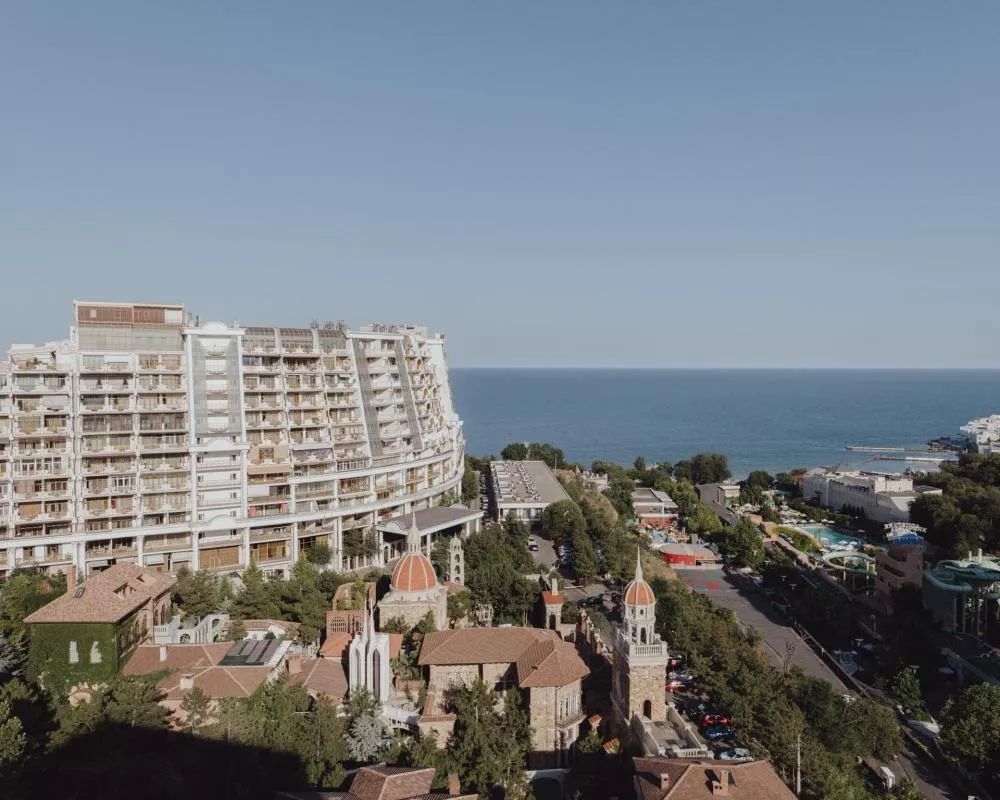 Un grand bâtiment résidentiel moderne surplombe la mer, entouré de bâtiments de style ancien avec des toits en tuiles rouges, le tout intégré dans un environnement verdoyant.