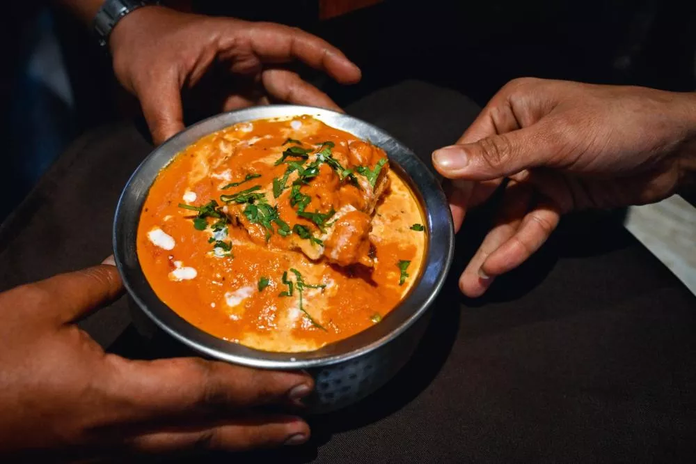 Un plat de curry crémeux, probablement du poulet au beurre, garni de coriandre fraîche, est tenu par deux mains.