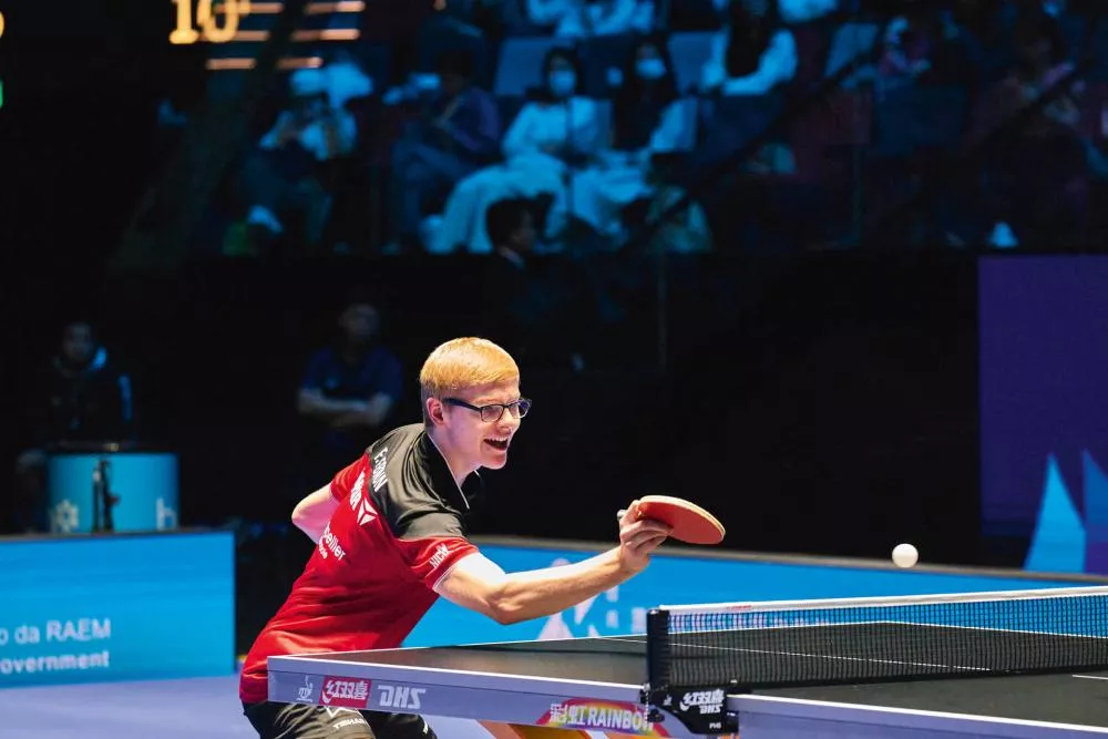 Un joueur de tennis de table en plein match, vêtu d'un maillot rouge et noir, prêt à frapper la balle avec sa raquette.