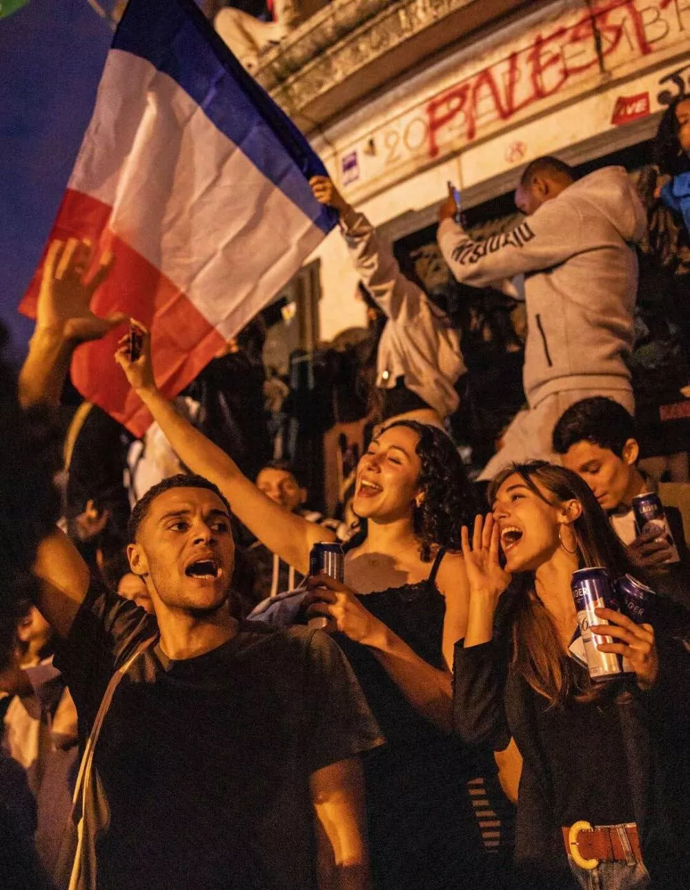 Des personnes célèbrent avec enthousiasme, brandissant des drapeaux français et des boissons, dans une ambiance festive et animée.