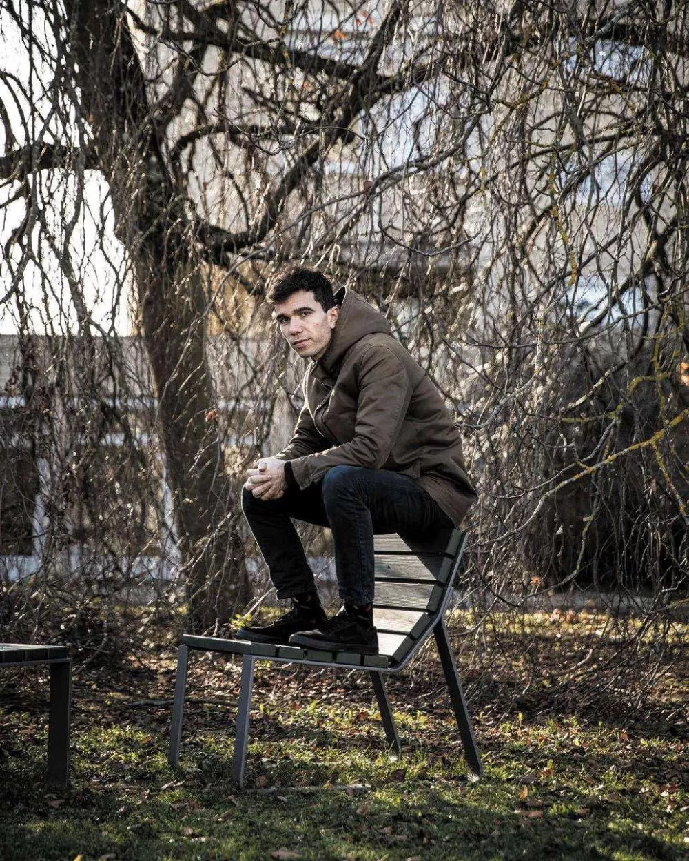 Un homme en veste marron est accroupi sur une chaise en plein air, entouré d'arbres aux branches dénudées.