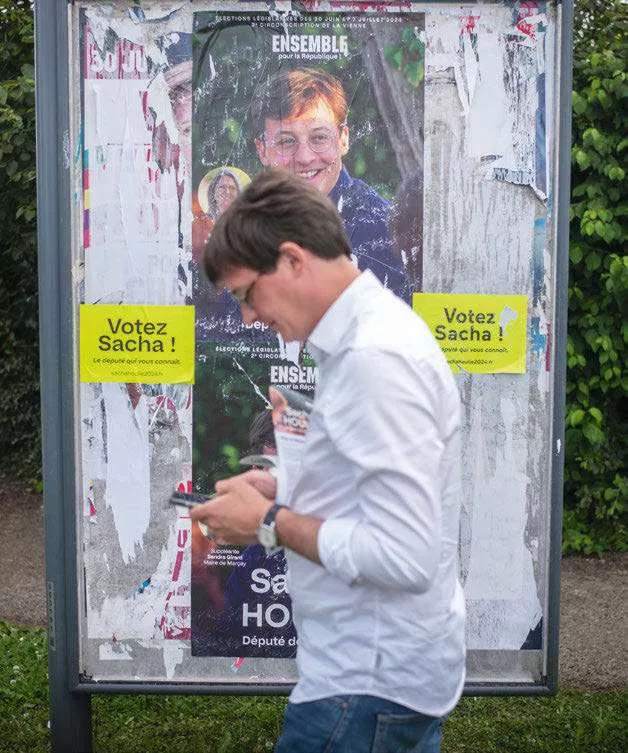 Une affiche électorale avec le slogan "Votez Sacha !" et un homme en chemise blanche marchant devant, regardant son téléphone.