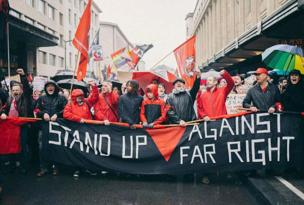Des personnes participent à une manifestation, tenant une banderole avec le texte "Stand Up Against Far Right". Ils portent des vêtements rouges et tiennent des drapeaux.