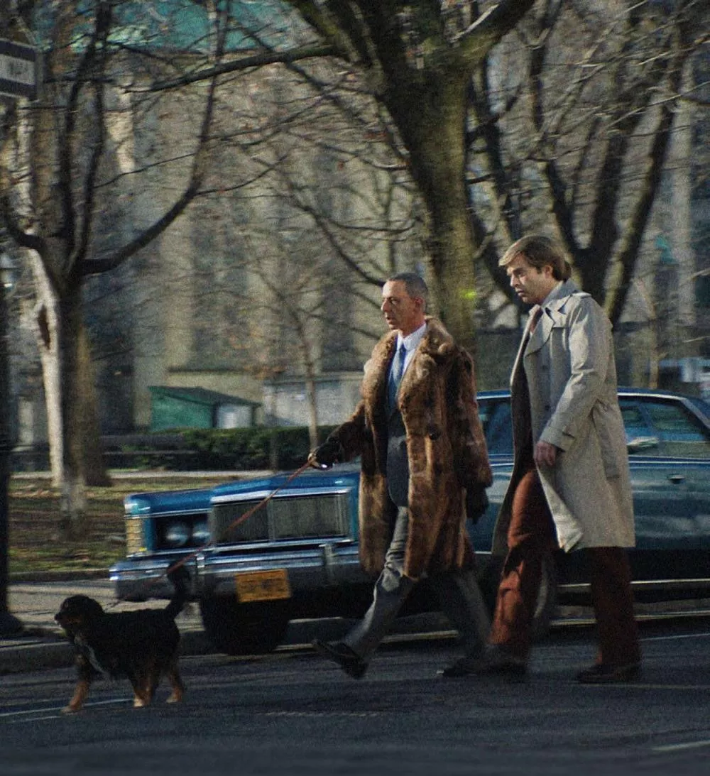 Deux hommes marchent dans la rue, l'un portant un manteau de fourrure et tenant un chien en laisse. Ils passent devant une voiture ancienne, et l'environnement est urbain avec des arbres dénudés.