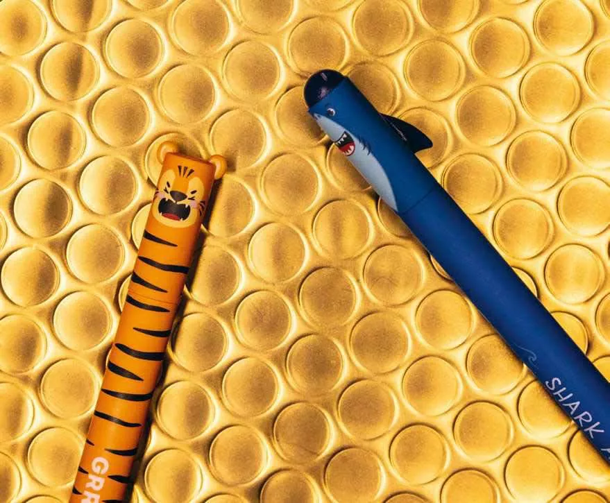 Deux stylos colorés sur un fond doré avec des motifs circulaires en relief. L'un est décoré comme un tigre et l'autre comme un requin.
