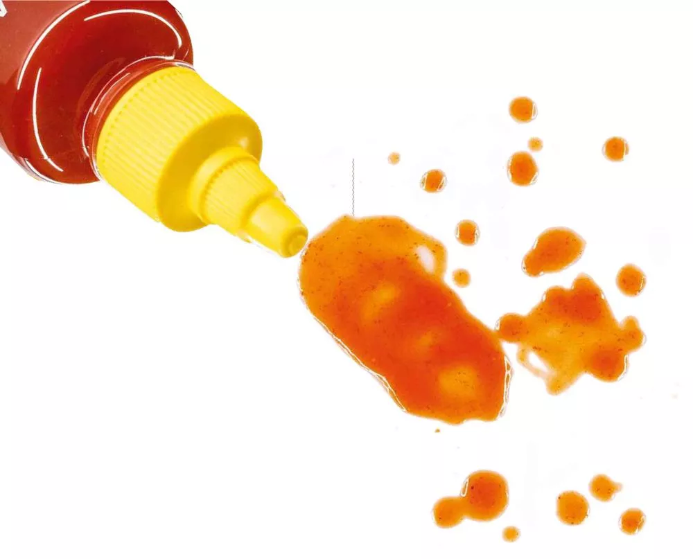 Une bouteille de sauce rouge avec un bouchon jaune verse de la sauce sur une surface blanche, créant des éclaboussures.