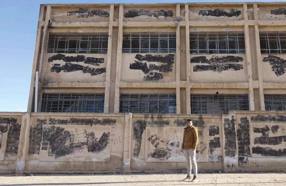 Un homme en manteau marron se tient devant un bâtiment en béton avec des fenêtres grillagées. Les murs du bâtiment et le mur d'enceinte sont marqués de taches sombres et de graffitis. Le ciel est dégagé.