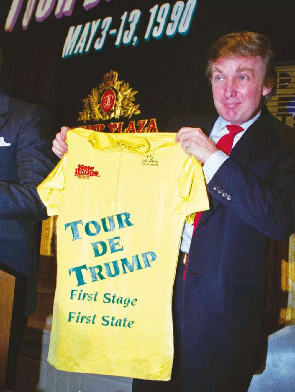 Un homme tient un maillot jaune avec l'inscription "TOUR DE TRUMP First Stage First State". En arrière-plan, une affiche indique "MAY 3-13, 1990".
