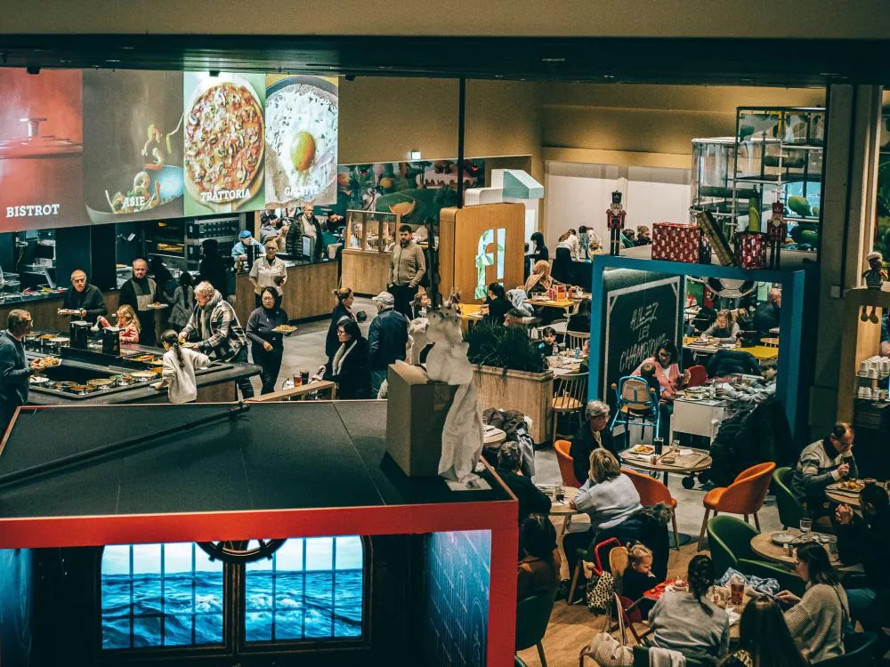 L'image montre un espace de restauration animé avec de nombreuses personnes assises à des tables, dégustant des repas. Il y a plusieurs stands de nourriture, chacun proposant différents types de cuisine comme "Bistrot", "Asie", "Trattoria" et "Galette". L'ambiance est conviviale et chaleureuse.