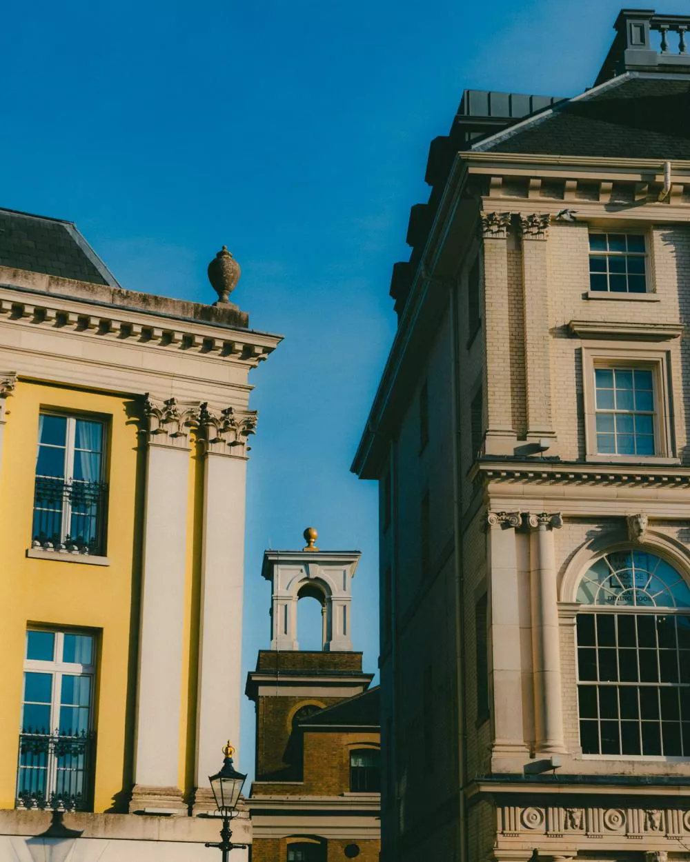 L'image montre des façades de bâtiments historiques avec des détails architecturaux classiques, des colonnes et des fenêtres à carreaux, sous un ciel bleu clair.