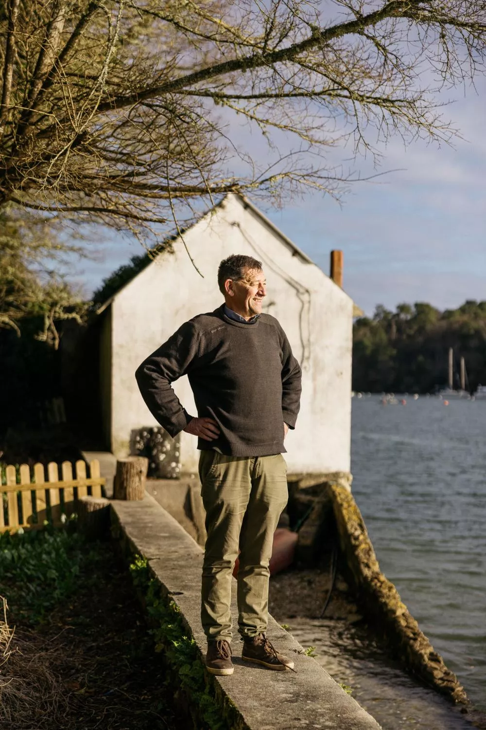 Un homme se tient debout près de l'eau, les mains sur les hanches, devant un bâtiment blanc. Il est habillé en pull foncé et pantalon kaki. L'environnement est naturel avec des arbres et un ciel dégagé.