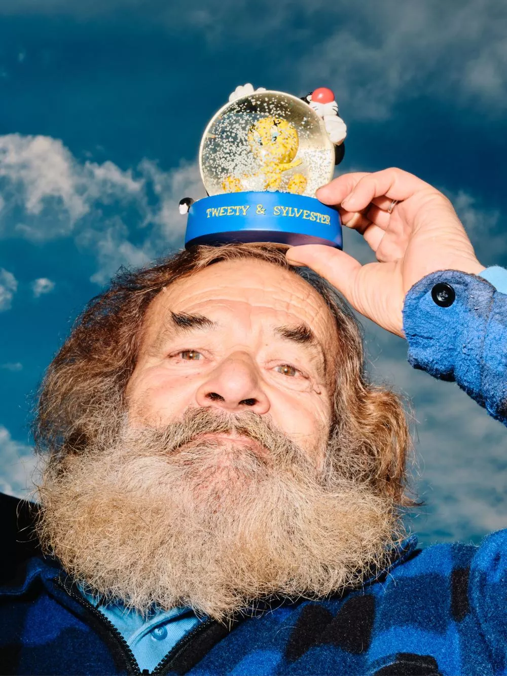 Un homme barbu tient une boule à neige "Tweety & Sylvester" sur sa tête, avec un ciel bleu en arrière-plan.