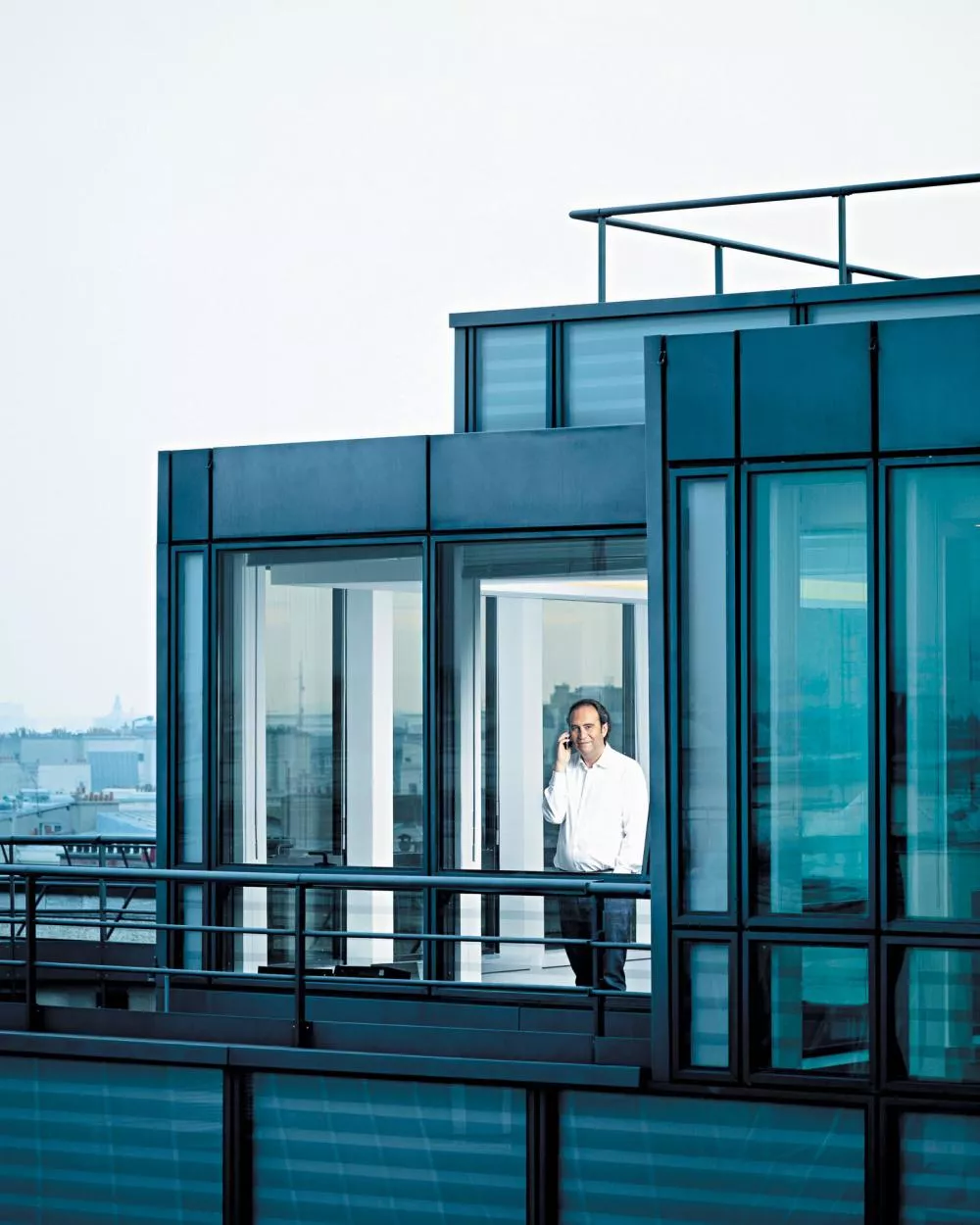 Un homme en chemise blanche se tient debout à l'intérieur d'un bâtiment moderne avec de grandes fenêtres en verre, tenant un téléphone à son oreille.
