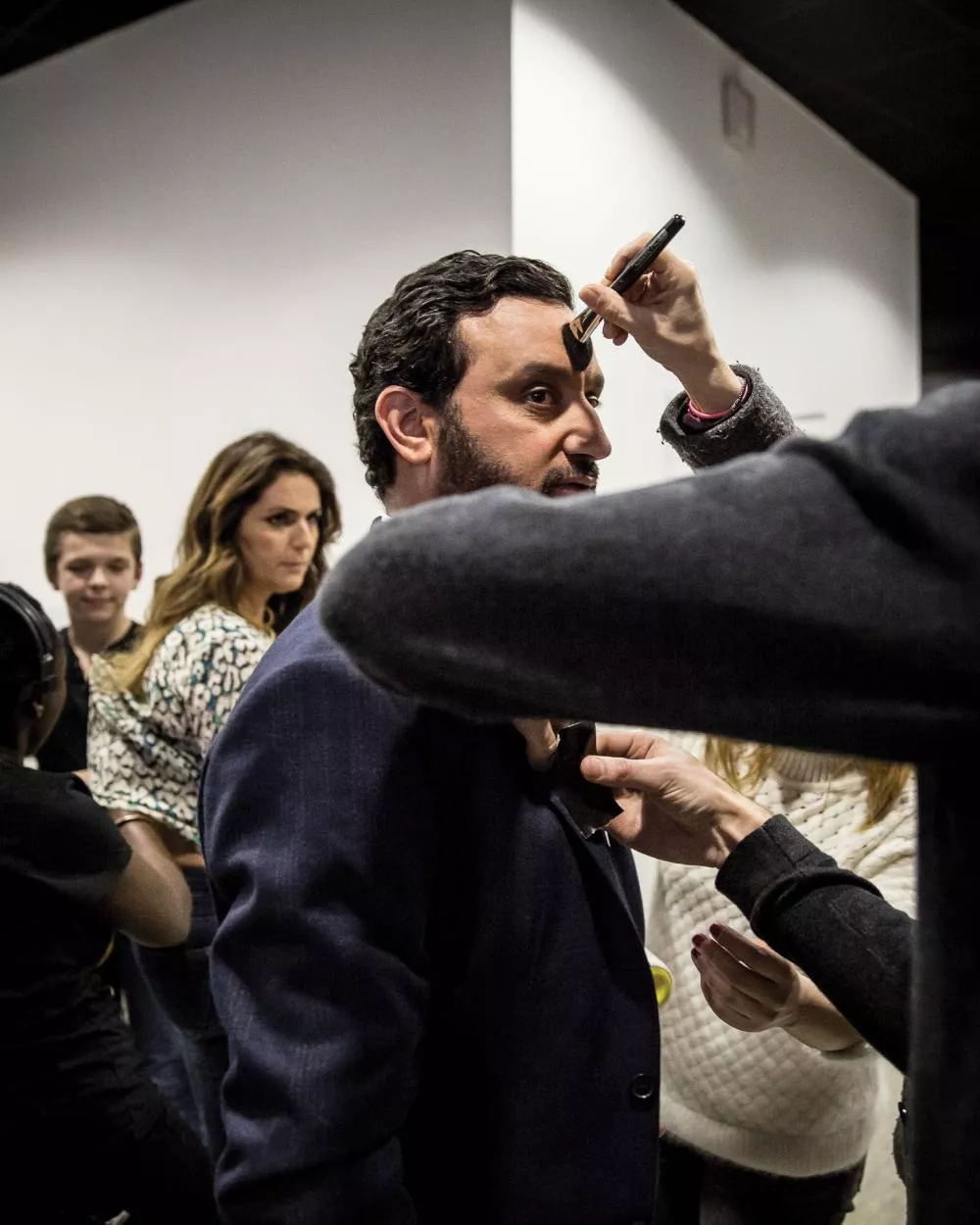 Un homme se fait maquiller et ajuster sa tenue par plusieurs personnes en coulisses, tandis que d'autres personnes se tiennent à l'arrière-plan.