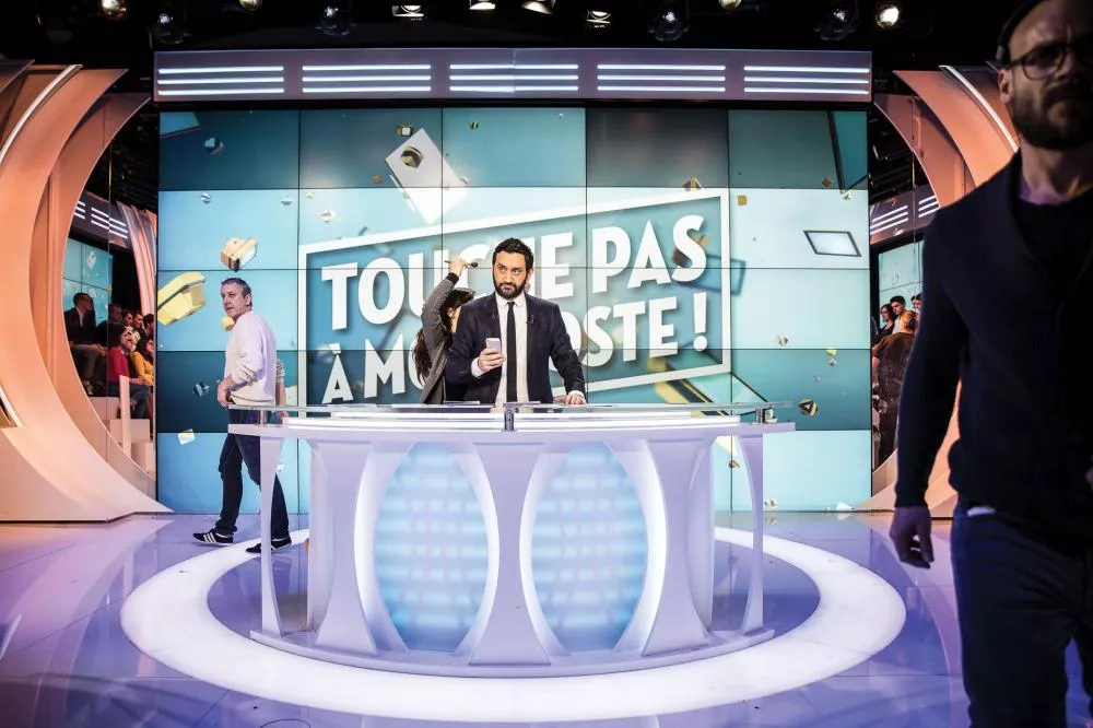 Une personne en costume est sur le plateau d'une émission de télévision avec le texte "TOUCHE PAS À MON POSTE !" affiché en arrière-plan. Plusieurs personnes se déplacent autour de lui.