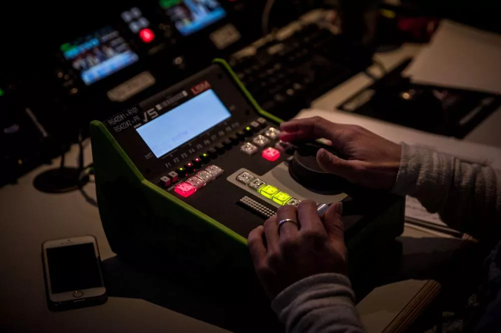 Une personne manipule une console de montage vidéo ou audio avec des boutons lumineux, dans un environnement sombre. Un téléphone est posé à côté de la console.