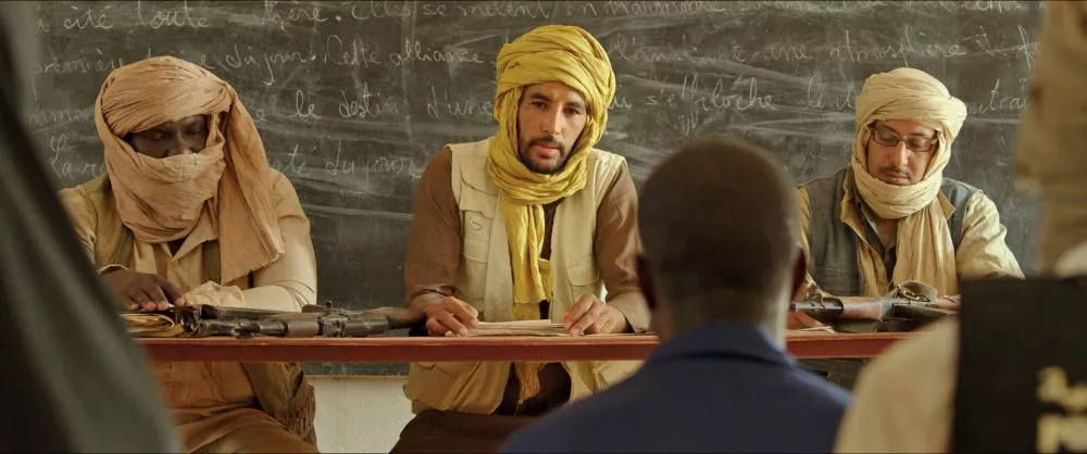 Trois hommes vêtus de turbans sont assis derrière une table avec des armes à feu posées devant eux. Ils semblent être dans une salle de classe, avec un tableau noir en arrière-plan où du texte est écrit à la craie. Un quatrième homme leur fait face, vu de dos.