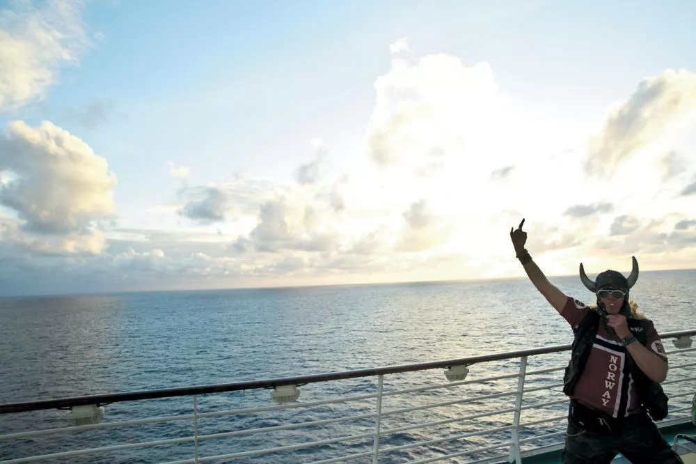 Un homme portant un casque à cornes et des lunettes de soleil se tient sur le pont d'un bateau, levant un bras en l'air. Il porte un t-shirt avec l'inscription "Norway". En arrière-plan, on voit la mer et un ciel partiellement nuageux.