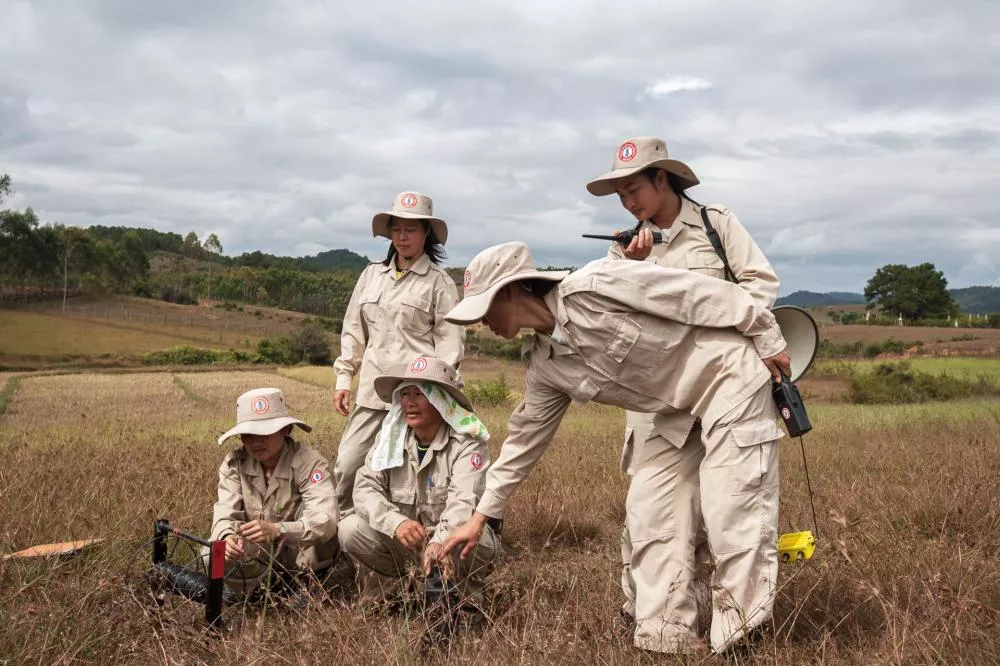Des personnes en uniforme beige et chapeau travaillent ensemble dans un champ. Elles semblent être engagées dans une activité de déminage ou d'exploration, utilisant des équipements spécialisés. Le paysage environnant est rural avec des collines et des arbres.