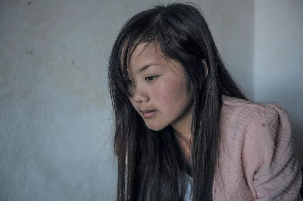 Une jeune femme aux longs cheveux noirs regarde vers le bas, vêtue d'une veste rose.