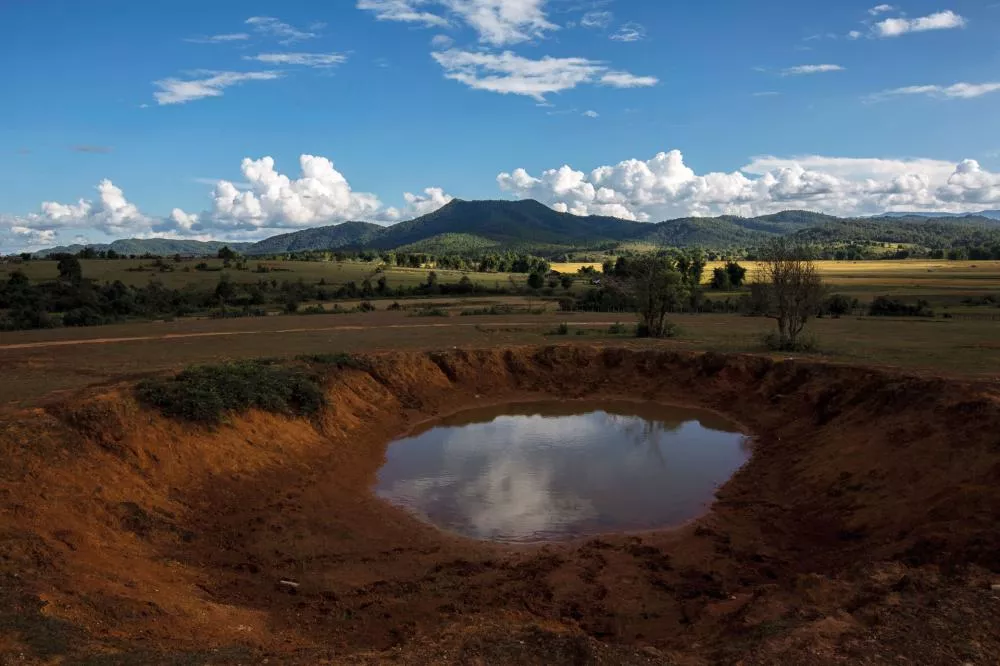 Un paysage naturel avec un petit plan d'eau entouré de terre rouge au premier plan, des prairies et des arbres au milieu, et des collines verdoyantes à l'horizon sous un ciel bleu avec des nuages blancs.