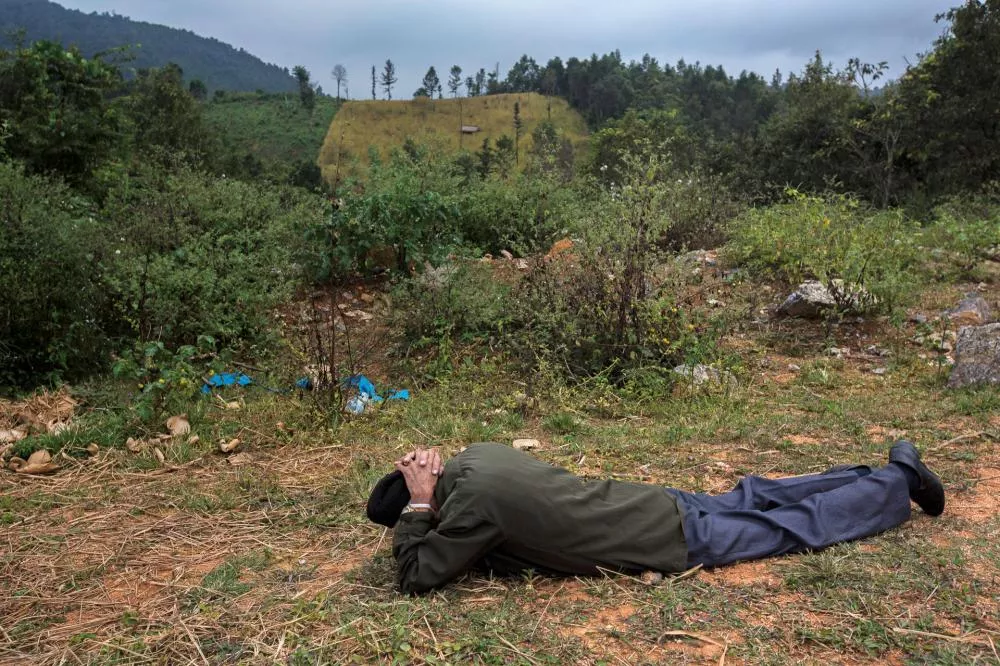 Un homme est allongé sur le sol, les mains croisées derrière la tête, dans un paysage naturel avec des collines et des arbres en arrière-plan.