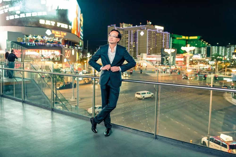 Un homme en costume se tient sur un balcon surplombant une rue animée avec des enseignes lumineuses et des bâtiments en arrière-plan, probablement à Las Vegas.