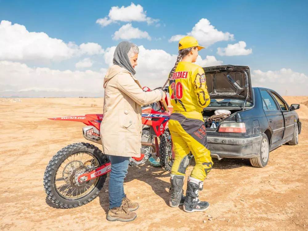 Une personne en tenue de motocross jaune se tient près d'une moto rouge, à côté d'une voiture avec le coffre ouvert dans un paysage désertique. Une autre personne, portant un manteau beige et un foulard, semble aider ou discuter avec elle.