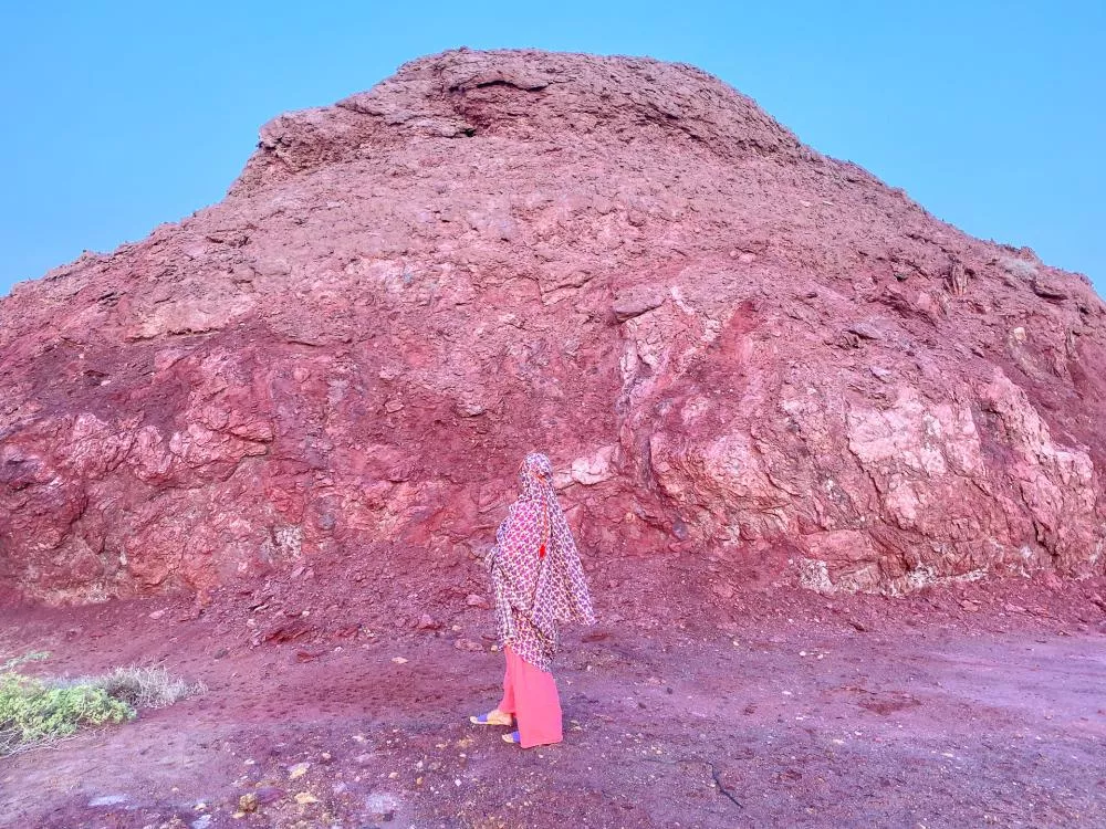 Une personne vêtue d'habits colorés se tient devant une grande formation rocheuse rougeâtre sous un ciel bleu.