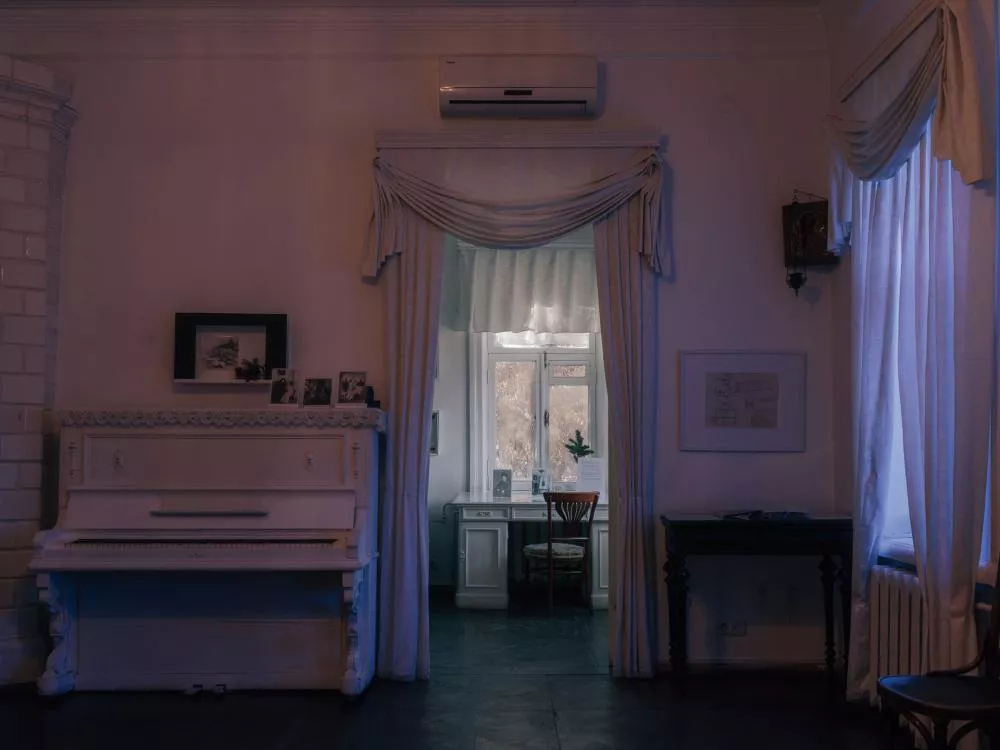 Une pièce intérieure avec un piano blanc à gauche, des rideaux élégants encadrant une ouverture vers une autre salle lumineuse, et des photos sur le piano.