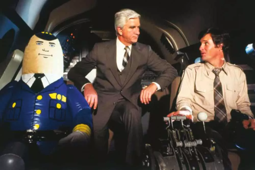 Un homme en costume est assis entre un pilote gonflable et un autre homme en chemise dans le cockpit d'un avion.