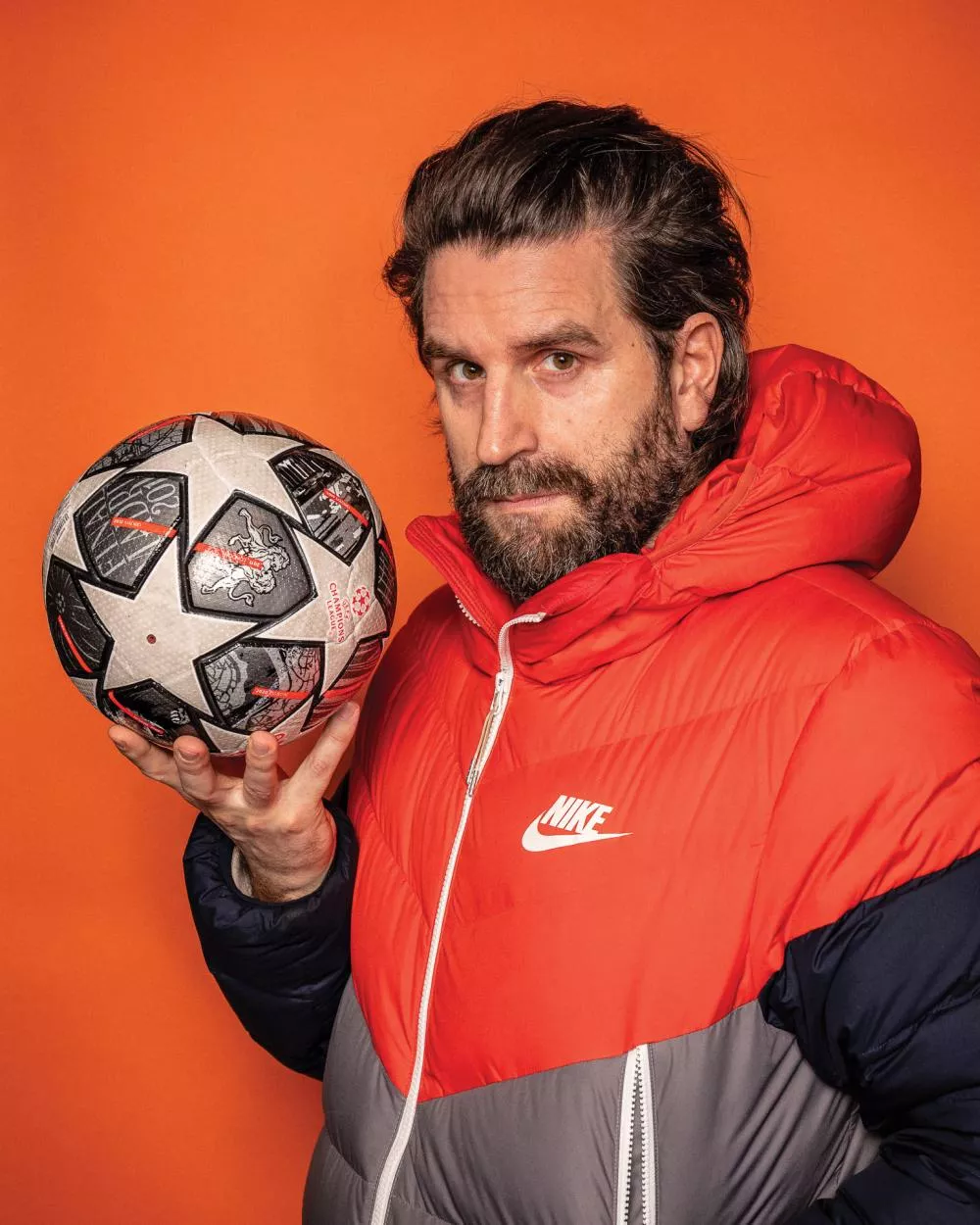 Un homme avec une barbe tient un ballon de football. Il porte une veste Nike rouge et grise avec une capuche. L'arrière-plan est orange.