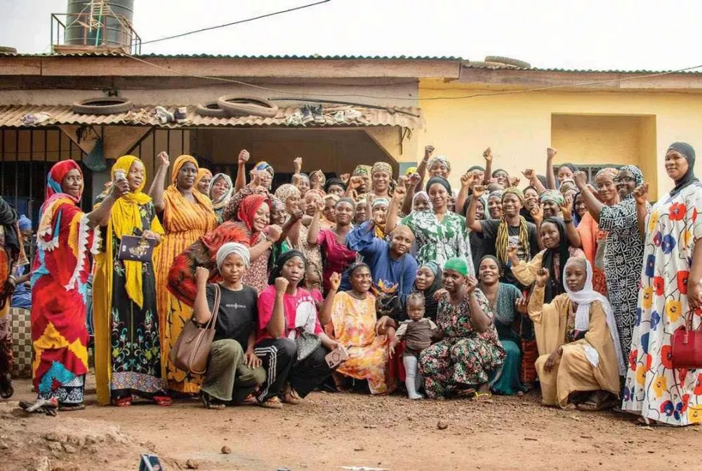 Un groupe de femmes et d'enfants pose ensemble devant un bâtiment, certaines levant le poing en signe de solidarité. Elles portent des vêtements colorés et semblent être dans un cadre communautaire ou de rassemblement.