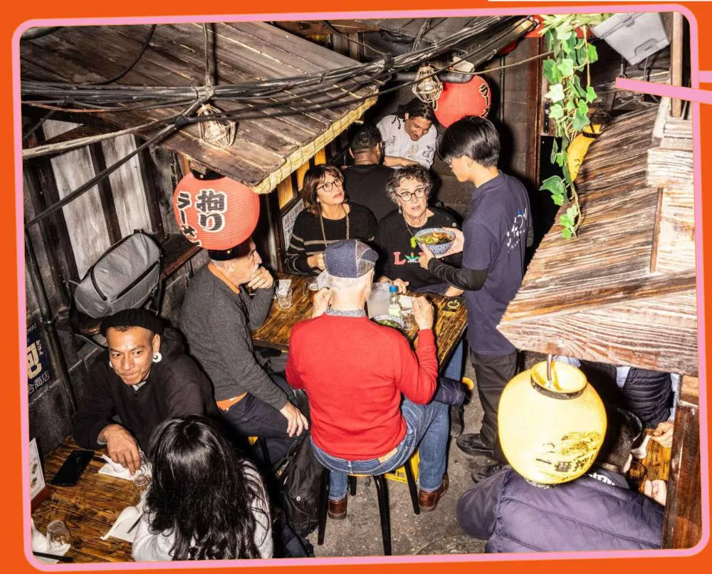 Des personnes sont assises autour de tables dans un restaurant animé, discutant et mangeant. Un serveur est debout, tenant un plat. L'ambiance est chaleureuse et conviviale, avec des lanternes japonaises décoratives.
