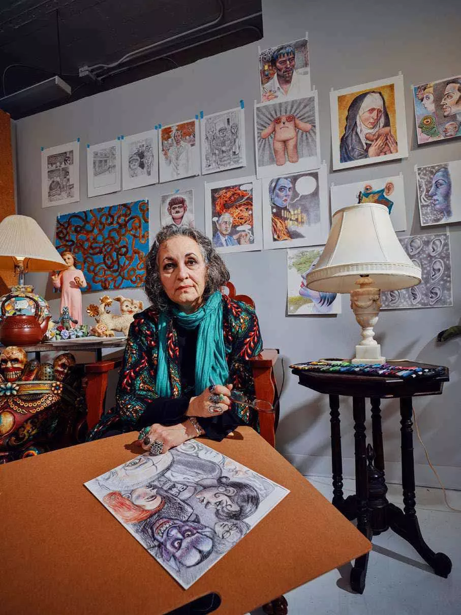 Une femme est assise dans une pièce remplie de dessins et d'œuvres d'art accrochés au mur. Elle porte un foulard turquoise et tient des lunettes. Devant elle, un dessin est posé sur une table. La pièce est décorée avec des objets et des lampes.