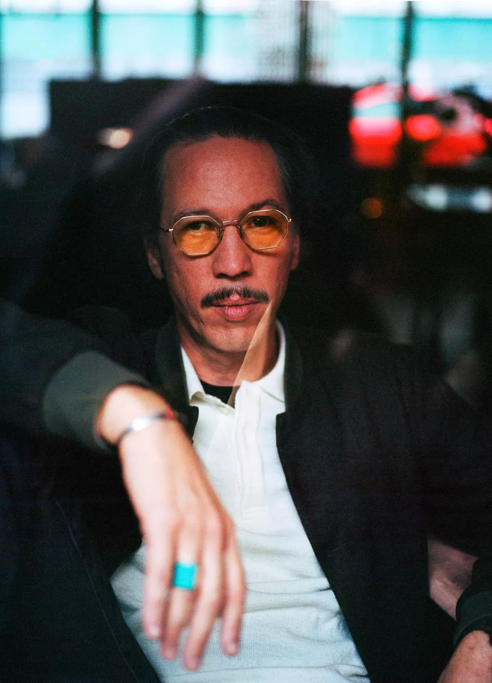 Un homme portant des lunettes teintées jaunes et une moustache est assis, vêtu d'une veste sombre et d'un polo clair. Il a une bague turquoise à la main. L'arrière-plan est flou avec des teintes rouges et vertes.