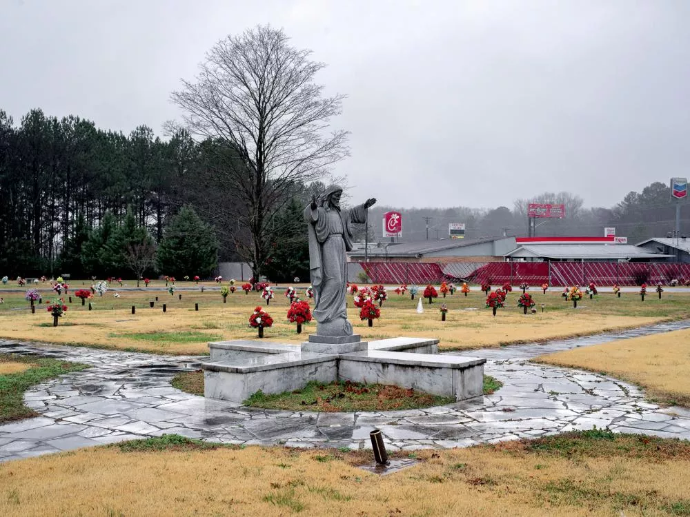Un cimetière avec une statue au centre, entouré de nombreuses tombes ornées de fleurs colorées. À l'arrière-plan, on aperçoit des bâtiments commerciaux et des panneaux publicitaires.