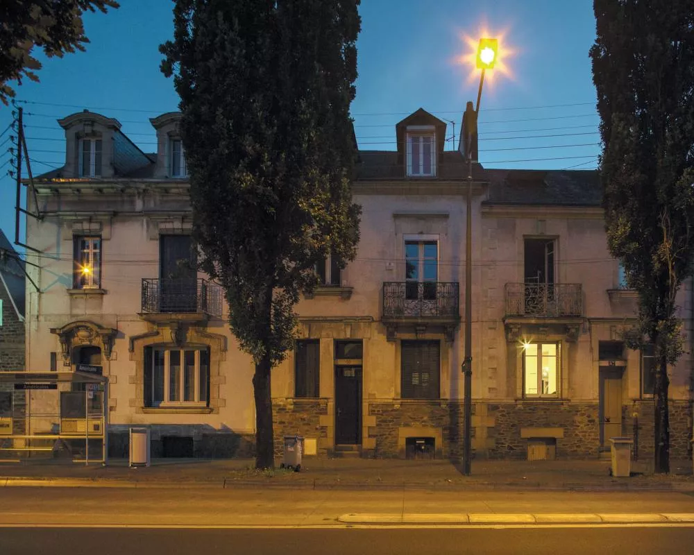 Une rue au crépuscule avec un bâtiment résidentiel ancien, des arbres et un lampadaire allumé.