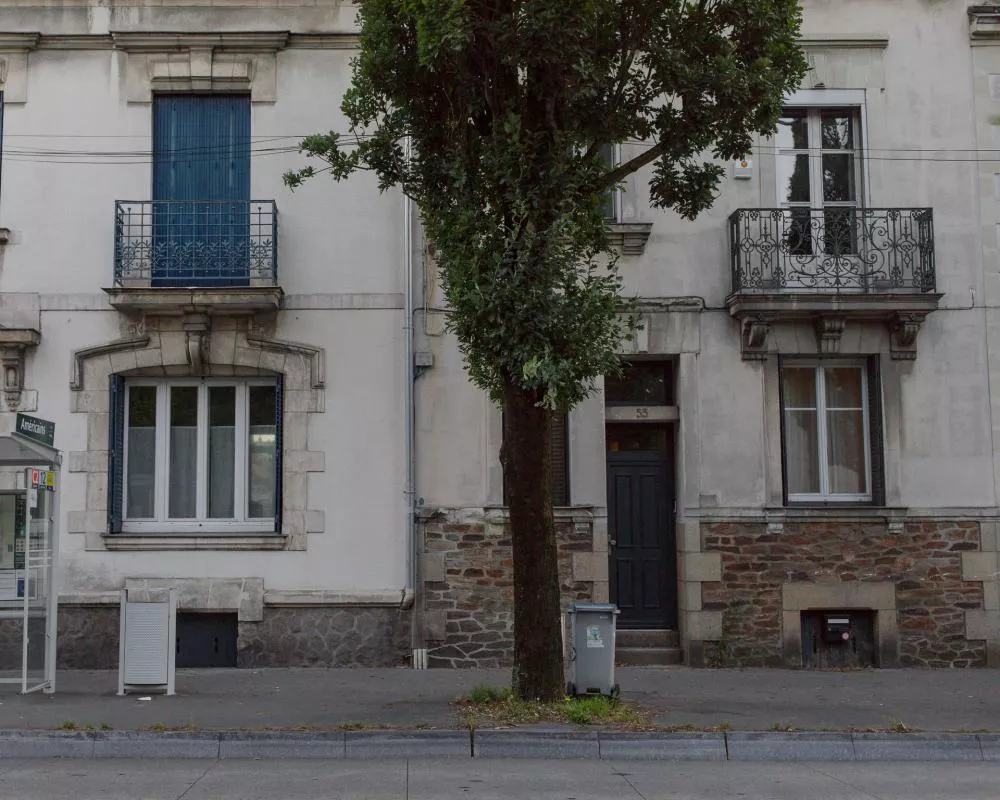 Une façade de bâtiment avec des balcons en fer forgé, des fenêtres et une porte d'entrée. Un arbre est situé devant le bâtiment, et une poubelle est visible sur le trottoir. À gauche, il y a un abribus avec le nom "Américains".
